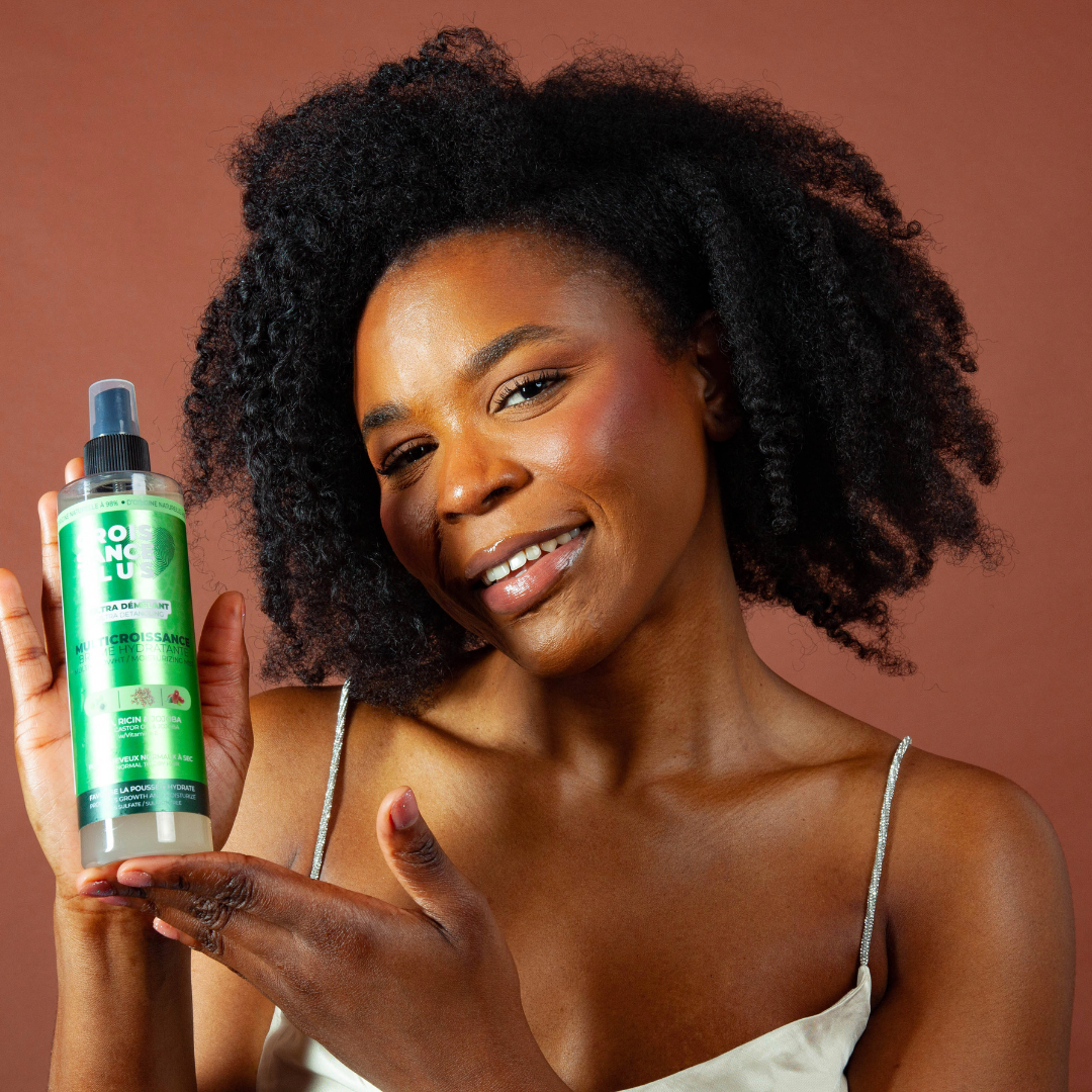 Smiling Black woman with natural curly hair holding a green bottle of Croissance Plus - Multricroissance Brume Hydratante 300ml Hair mist product by JS Organics