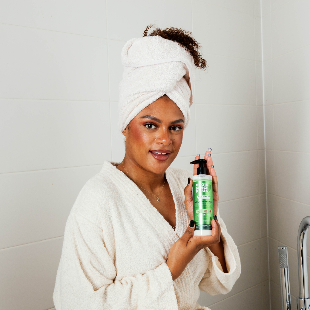 A smiling woman wearing a white robe and towel turban holds a pump bottle of Croissance Plus Multricroissance Lait Coiffant 300ml hair growth milk by JS Organics