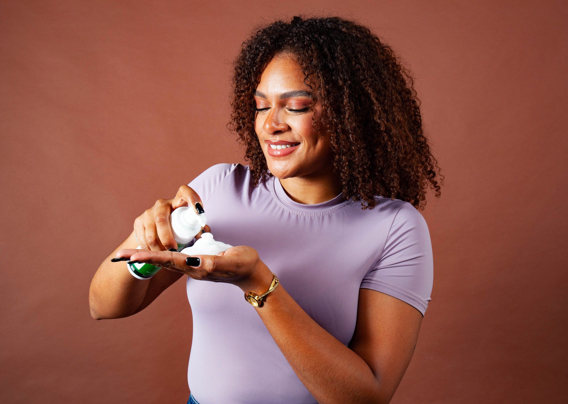 Smiling woman with curly hair dispensing Croissance Plus Mousse Coiffante Amla hair mousse by JS Organics into her hand.