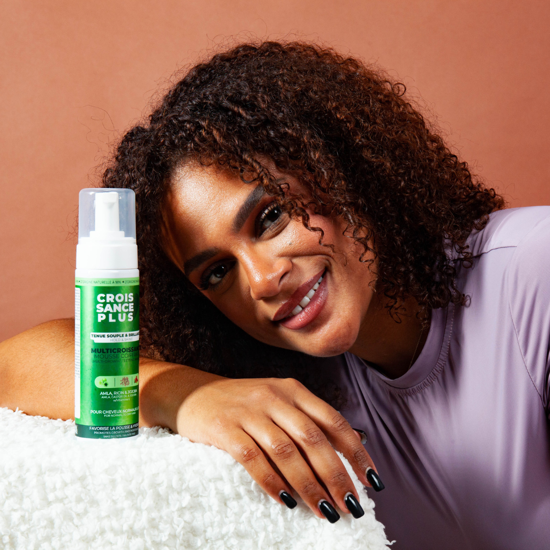 A smiling woman with a bottle of Croissance Plus Multricroissance Mousse Coiffante 200ml by JS Organics, which is a hair styling product.