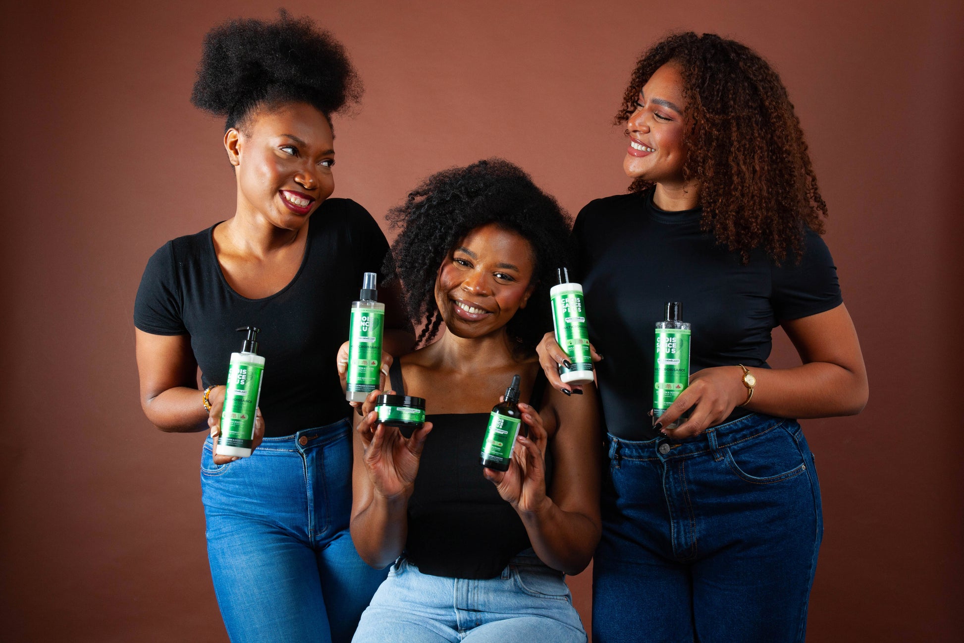 Three smiling Black women holding green and white Croissance Plus hair care products bottles by JS Organics.